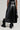 A person wearing the KEEP WALKING MIDI SKIRT, paired with black combat boots, stands on a light gray background. They are holding a black textured handbag, showcasing an outfit that beautifully blends edgy and chic styles.