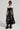 Dressed in the chic KEEP WALKING MIDI SKIRT, a woman showcases her confidence in an ensemble featuring a white top with black accents, paired with unique baggy boots and high-rise sunglasses. She poses effortlessly against a plain background, exuding style and poise.
