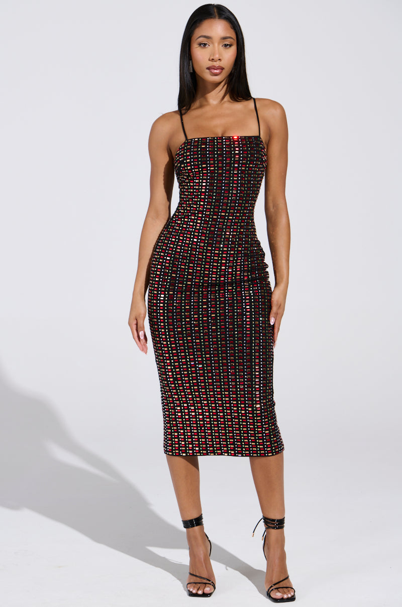 A woman stands confidently in the RENEE RHINESTONE MIDI DRESS, a bodycon fit with a sleeveless design and colorful dots, paired with black high-heeled sandals. The plain background emphasizes her silhouette and shadow.
