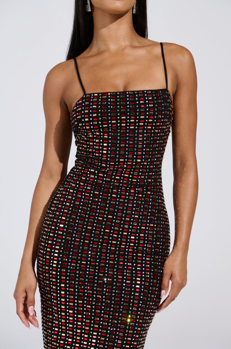 A person wears the RENEE RHINESTONE MIDI DRESS, featuring a sleeveless design with thin straps. It has a pattern of glittering red and silver squares on a black background. The cotton-blend bodycon fit accentuates their figure, complementing their visible black hair.