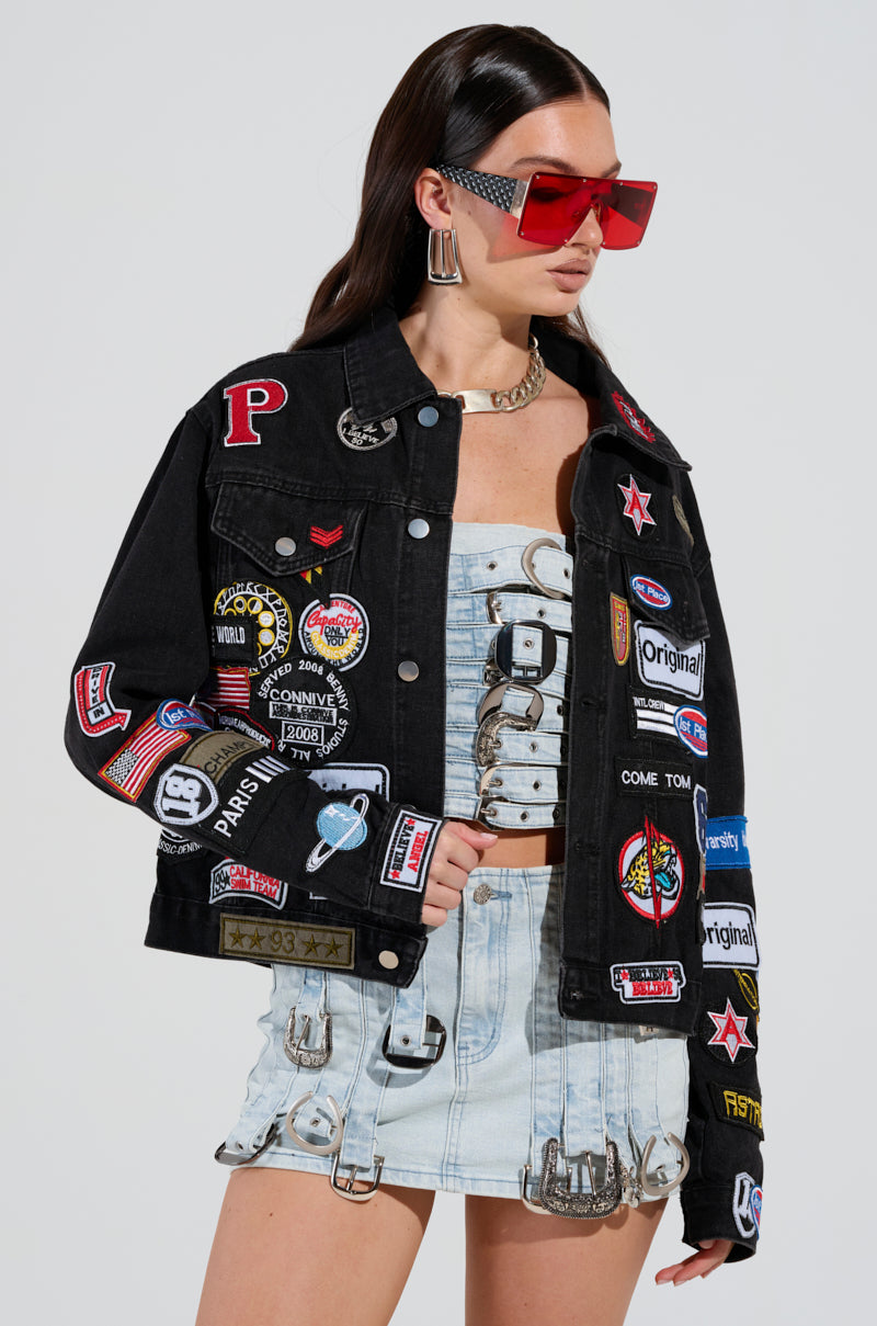 A person wearing the PATCH IT UP DENIM JACKET, which is cropped and embellished with vibrant patches reminiscent of a letterman jacket, over a light blue denim ensemble featuring metal buckles. They have long dark hair and accessorize with oversized red sunglasses and a chunky necklace.