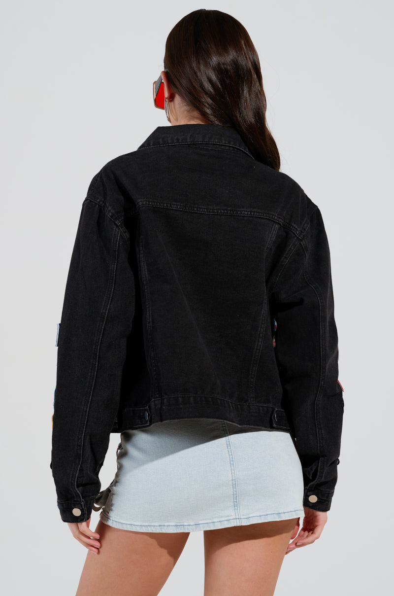A person with long dark hair in a cropped style is facing away, wearing the PATCH IT UP DENIM JACKET—a black denim piece decorated with letterman patches—and a light blue denim mini skirt. Red sunglasses sit atop their head against a neutral backdrop.