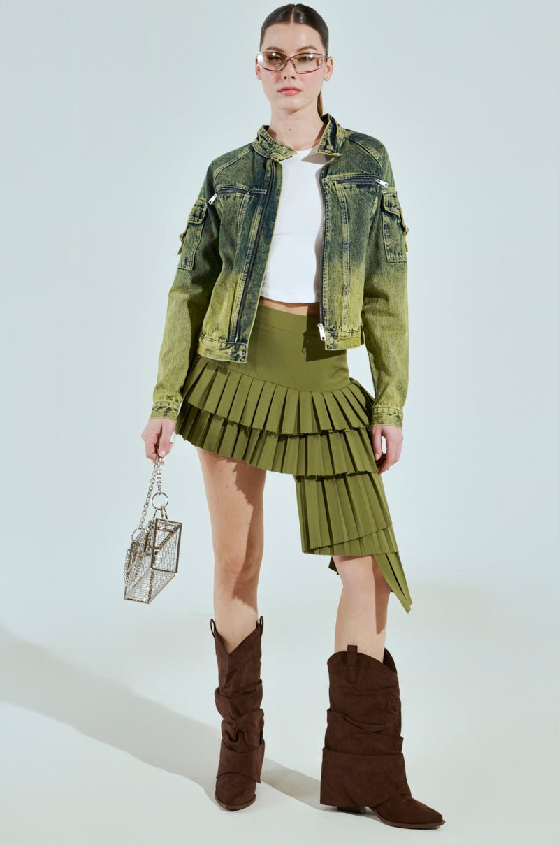 Someone wearing sunglasses sports the CALL ME CRAZY DENIM MOTO jacket, a white crop top, and an asymmetrical olive green pleated skirt. They carry a geometric handbag and wear brown slouchy boots against a light-colored background.