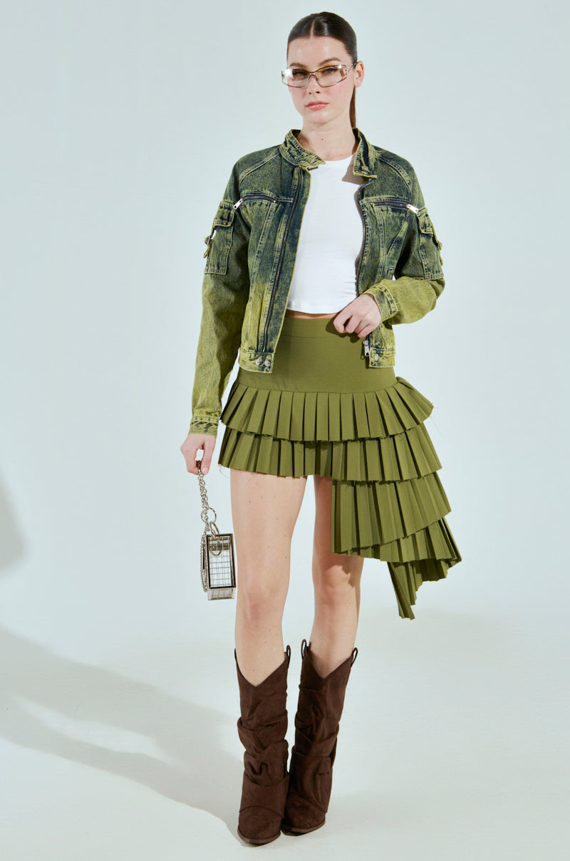 Wearing the CALL ME CRAZY DENIM MOTO jacket over a white top and asymmetrical olive green pleated skirt, they carry a small chain bag and wear brown boots. Their hair is tied back, glasses on their nose, achieving an effortlessly chic look.