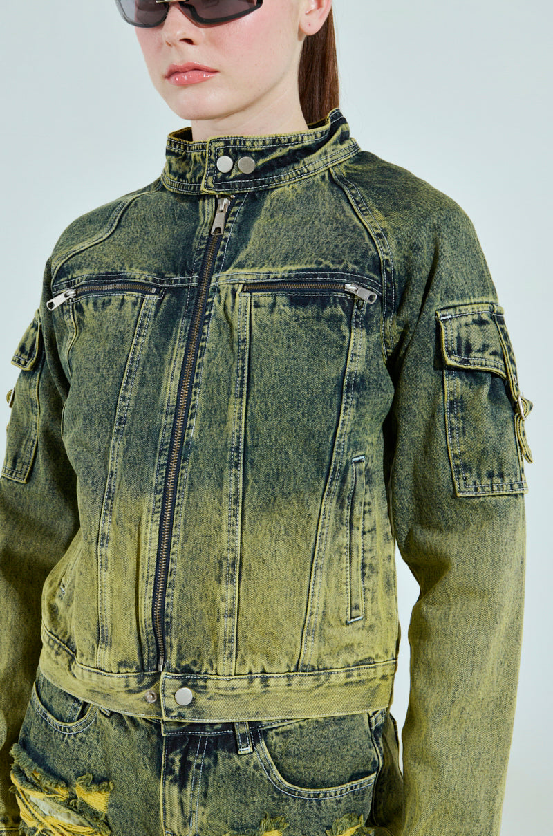 A person wearing sunglasses and the CALL ME CRAZY DENIM MOTO jacket, featuring a front zipper, multiple pockets, and a yellow-green two-tone colorway. Its cropped silhouette adds flair against a plain background, making the ensemble stand out.