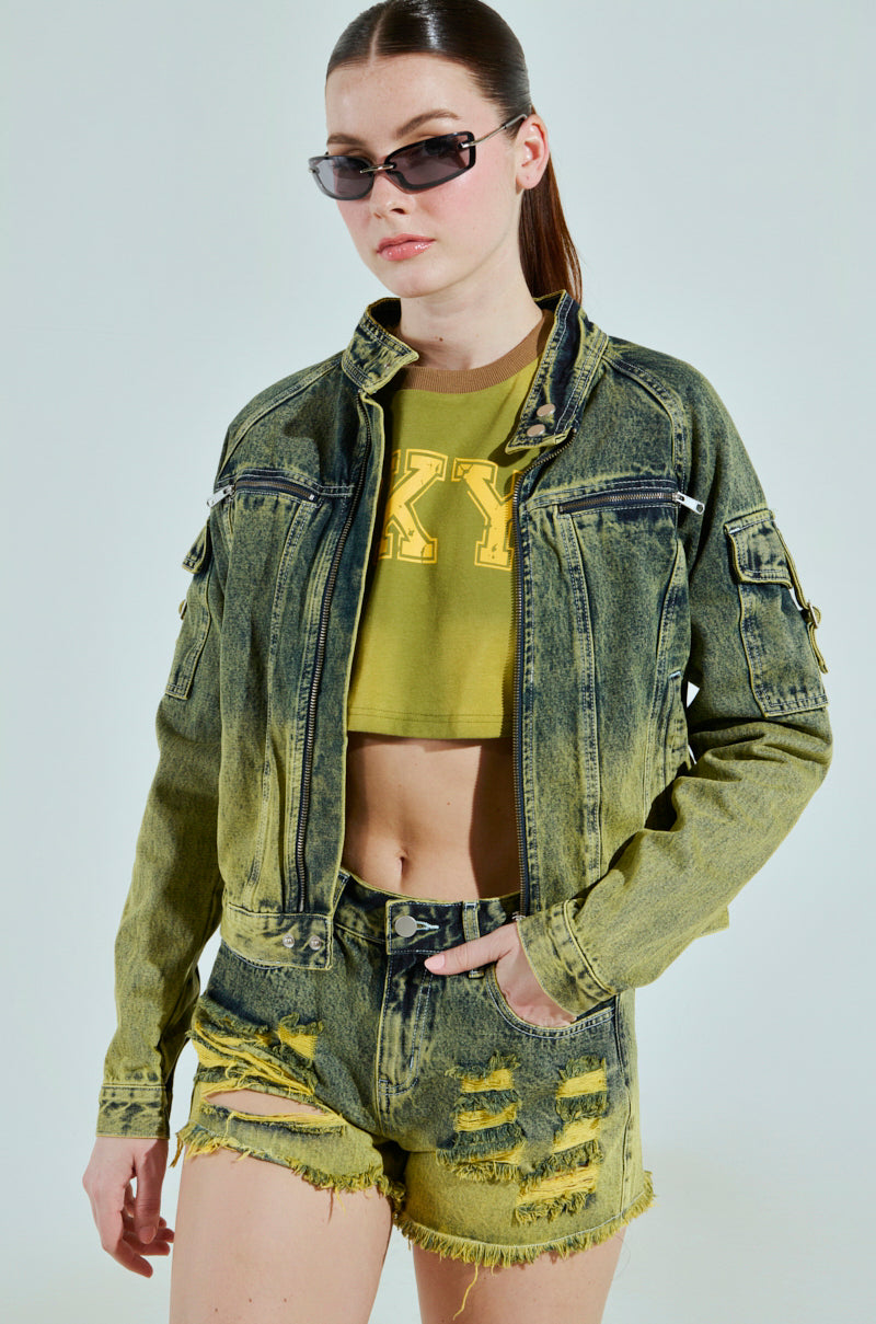 A person wearing dark sunglasses, the "Call Me Crazy Denim Moto" jacket, a yellow crop top with bold lettering, and distressed shorts poses against a plain background. The cropped silhouette features a two-tone colorway with frayed edges.