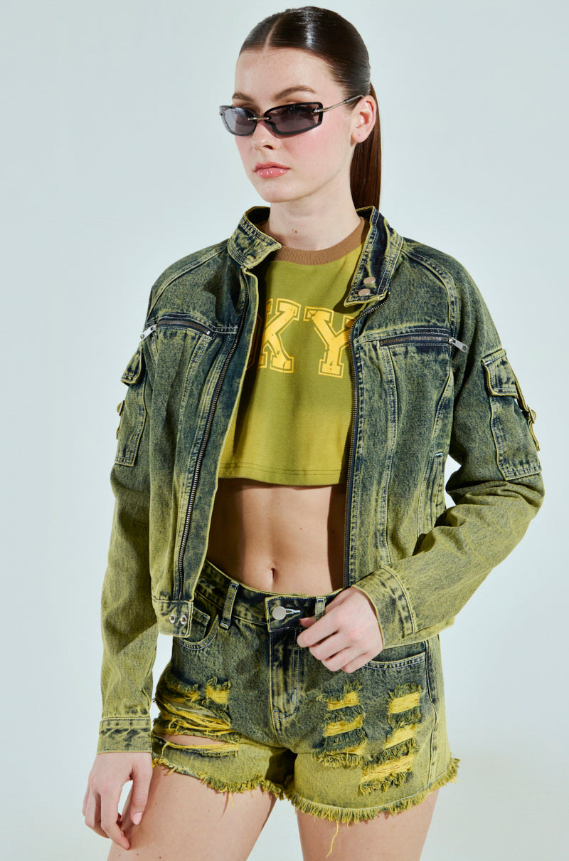 A person with long hair tied back, wearing dark sunglasses and a green cropped silhouette top, dons the CALL ME CRAZY DENIM MOTO jacket with frayed denim shorts. They stand against a plain background, exuding confidence in their two-tone colorway ensemble.