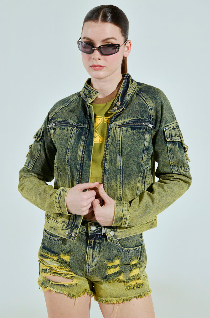A person in sunglasses poses in the CALL ME CRAZY DENIM MOTO, a green two-tone denim jacket with matching distressed shorts and a coordinating shirt. Their hair is tied back against a light backdrop.
