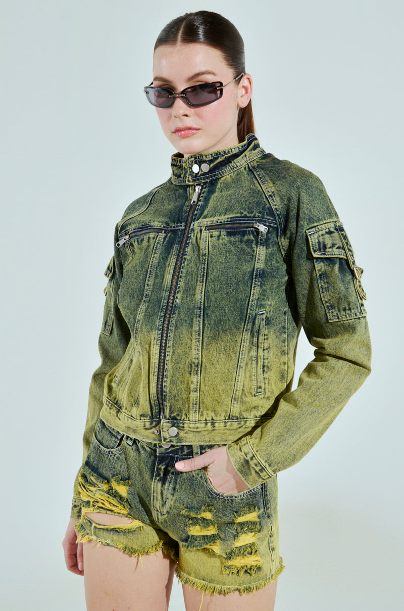 Someone in the CALL ME CRAZY DENIM MOTO jacket and matching frayed shorts exudes confidence. Their slicked-back hair and dark sunglasses enhance their edgy look against a light, neutral background, creating a striking silhouette.