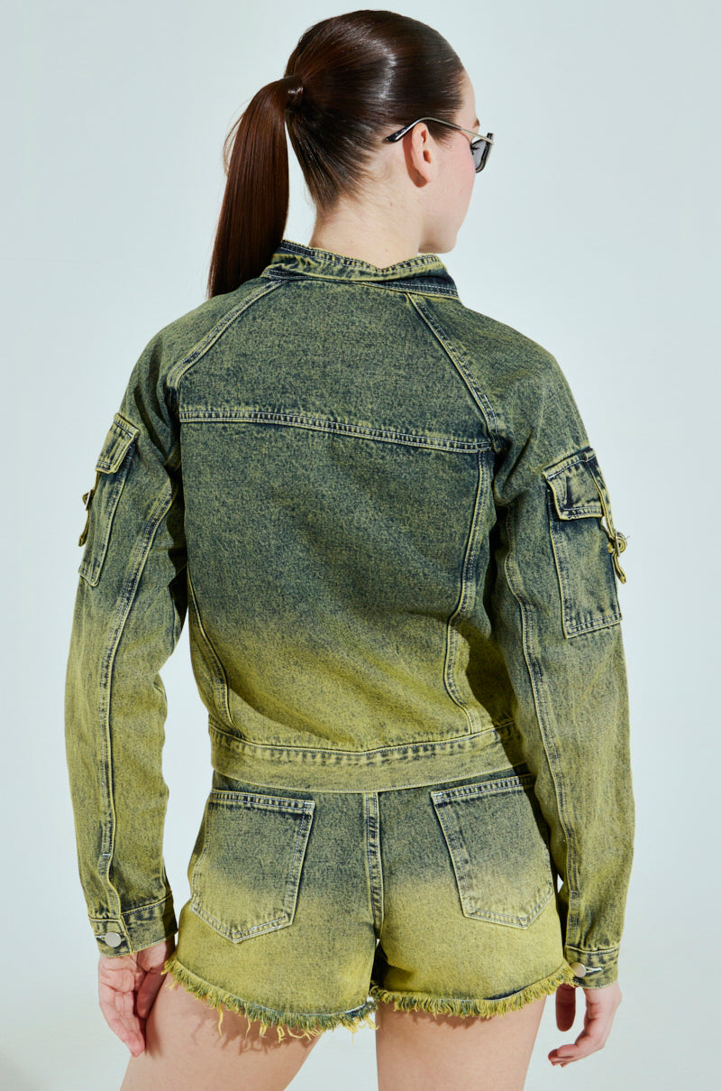 With long hair tied back, a person faces away from the camera, wearing the CALL ME CRAZY DENIM MOTO jacket and matching shorts in green and yellow. The outfit has a cropped silhouette and showcases a faded, distressed style.