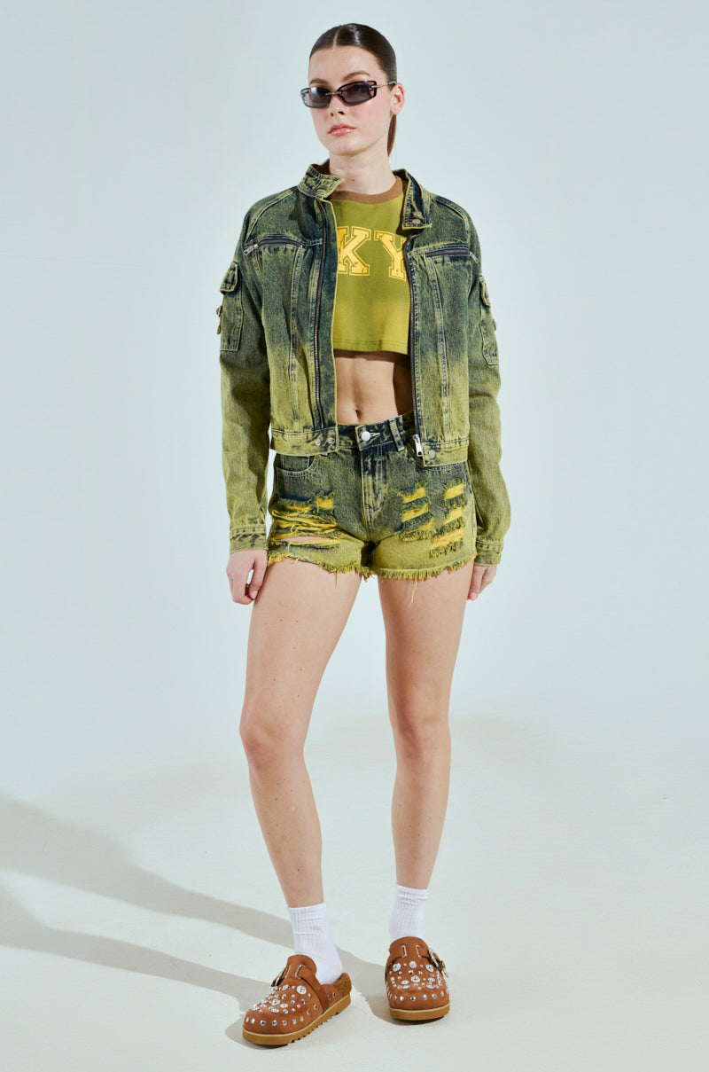 Modeling sunglasses, they sport the chic CALL ME CRAZY DENIM MOTO jacket and shorts set with a green crop top, brown studded slip-on shoes, and white socks against a plain light gray backdrop.