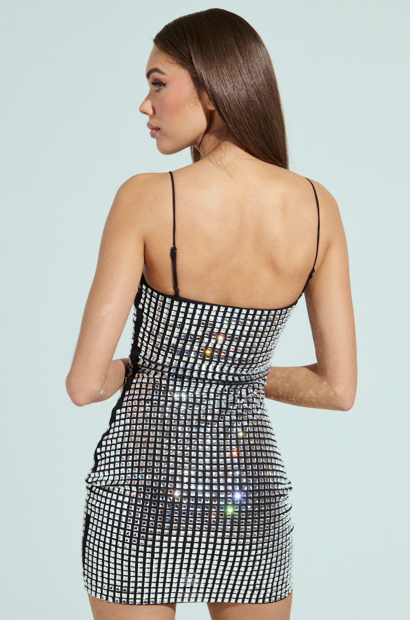 A woman with long brown hair stands facing away from the camera, highlighting the glimmering IN MY ELEMENT RHINESTONE MESH MINI DRESS. This sleeveless bodycon silhouette features reflective square sequins on a solid light background.