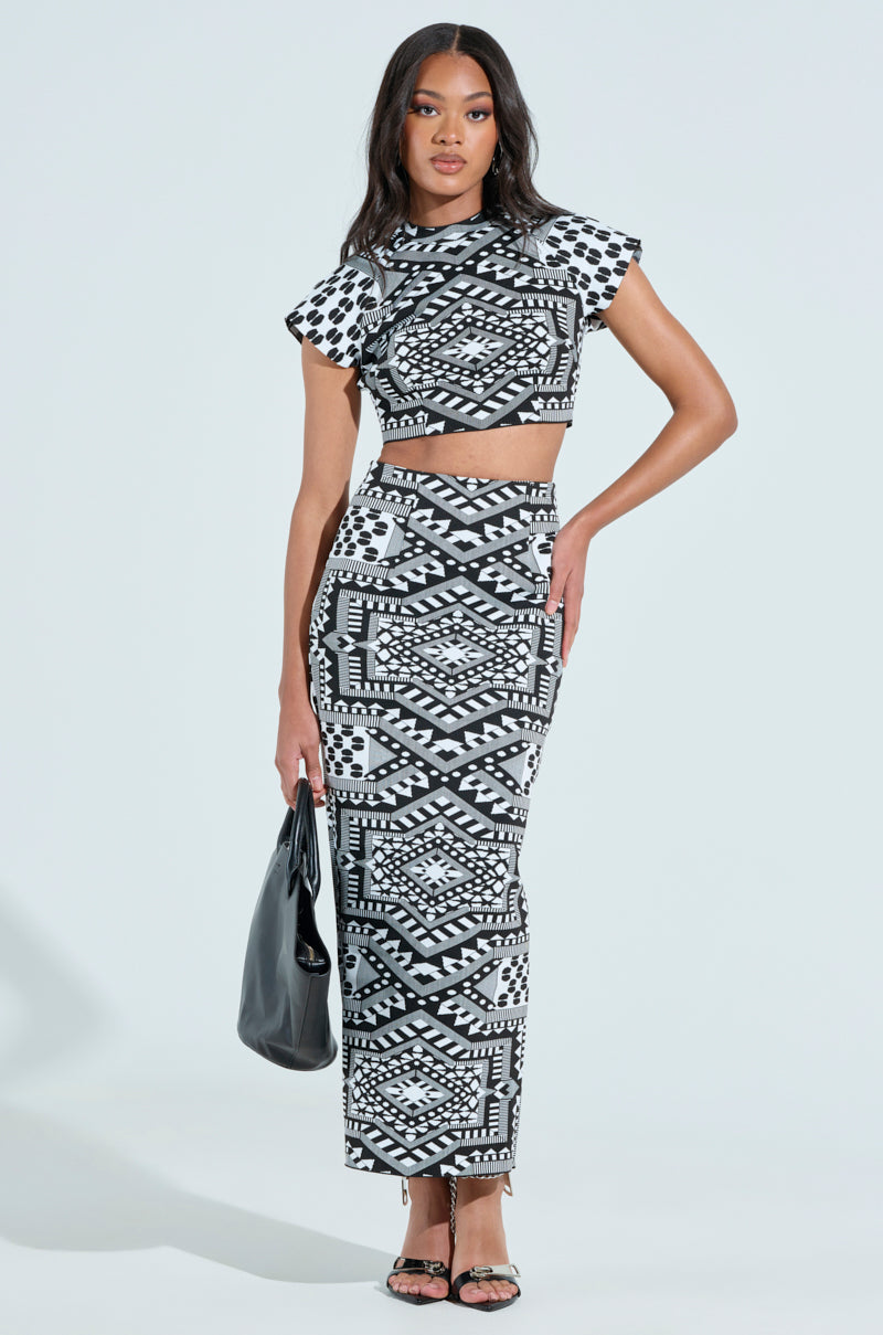 Someone is wearing a fashionable Vaca Mode Abstract Maxi Skirt featuring a geometric B&W pattern. It's paired with a crop top, a black handbag, and trendy strappy high heels against a plain light background, emphasizing the outfit's bold high-rise design.