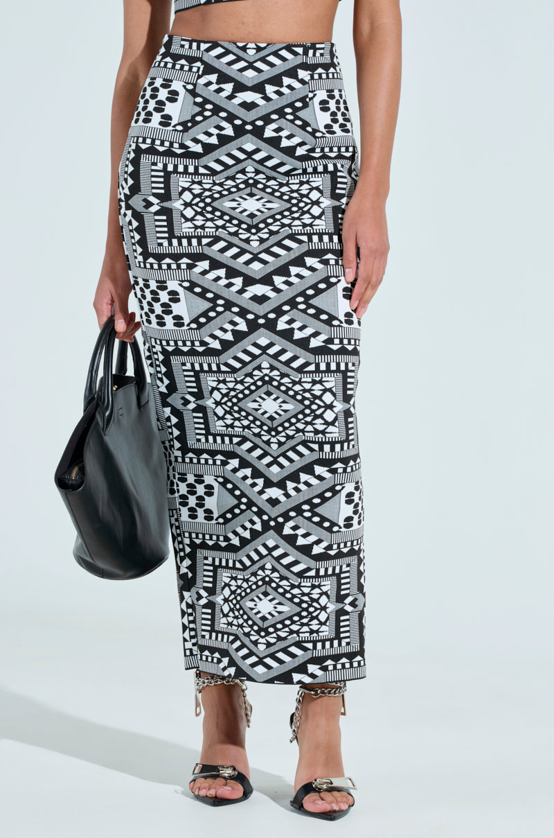 Someone is wearing the VACA MODE ABSTRACT MAXI SKIRT with a bold geometric B&W pattern. They carry a black handbag and wear black high-heeled sandals. The plain, light background highlights the skirt's striking design.