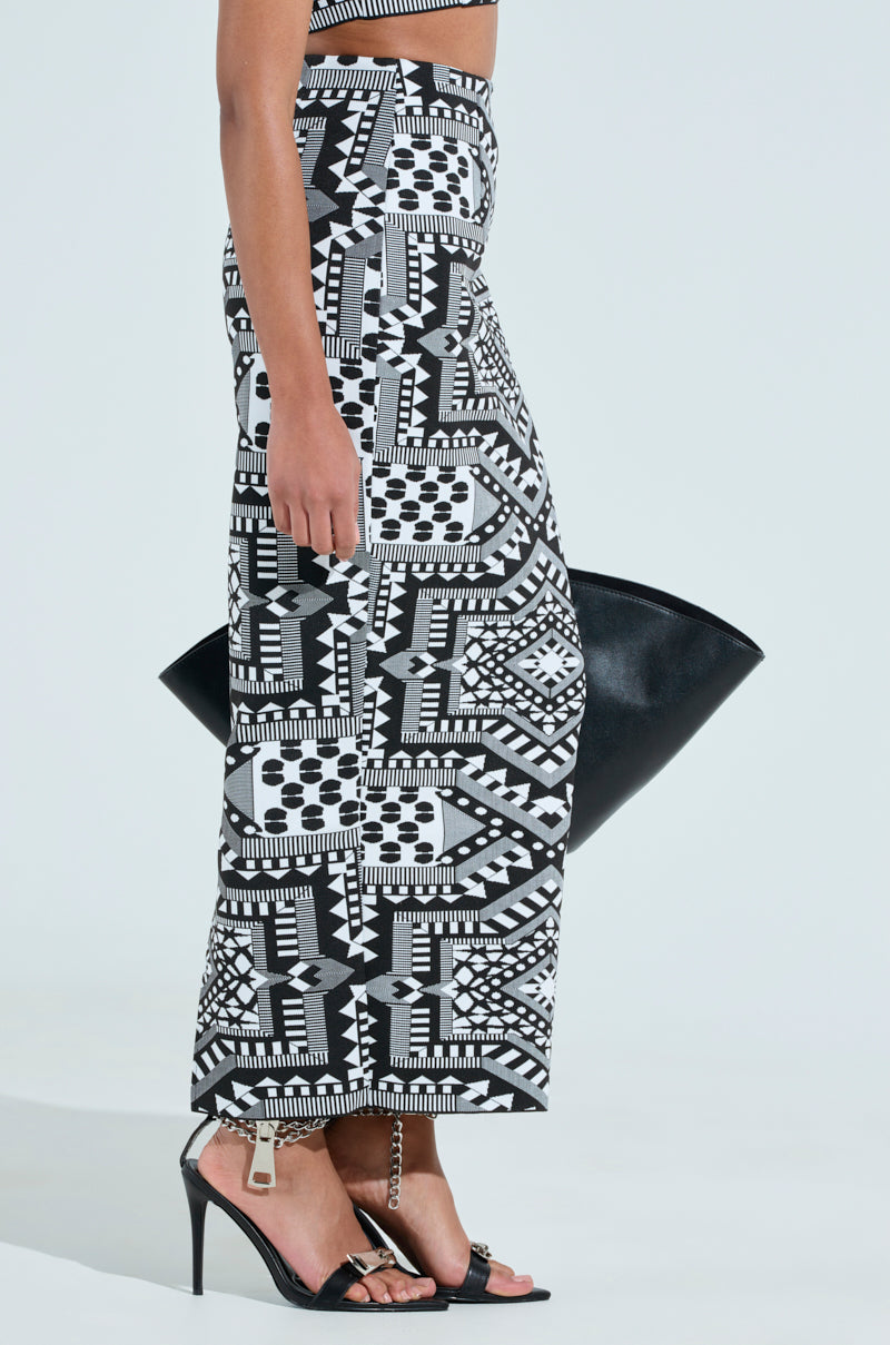 A person wearing a VACA MODE ABSTRACT MAXI SKIRT with a geometric black-and-white design pairs it with black high-heeled sandals and holds a large black handbag. The stylish look is captured from the side against a light background.
