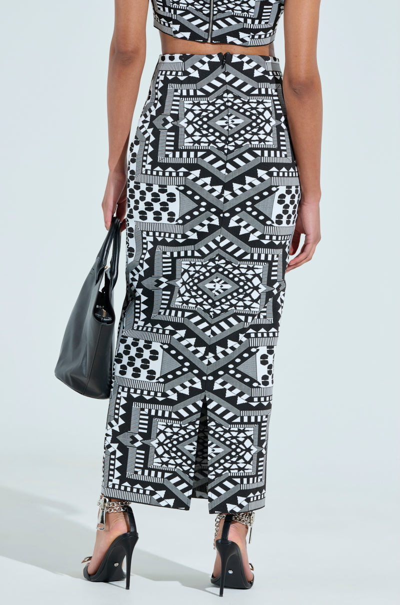 Rear view of a person in a black and white geometric outfit with the "Vaca Mode Abstract Maxi Skirt" featuring a high-rise design and back slit, paired with a matching crop top. They carry a black handbag and wear high-heeled sandals.