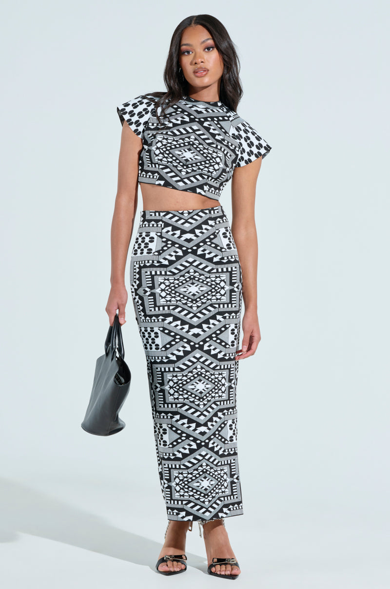 A woman confidently wears a VACA MODE Abstract Maxi Skirt and matching geometric B&W crop top. She carries a black handbag, paired with black strappy sandals. Her long hair cascades freely against a plain background, highlighting her stylish high-rise ensemble.