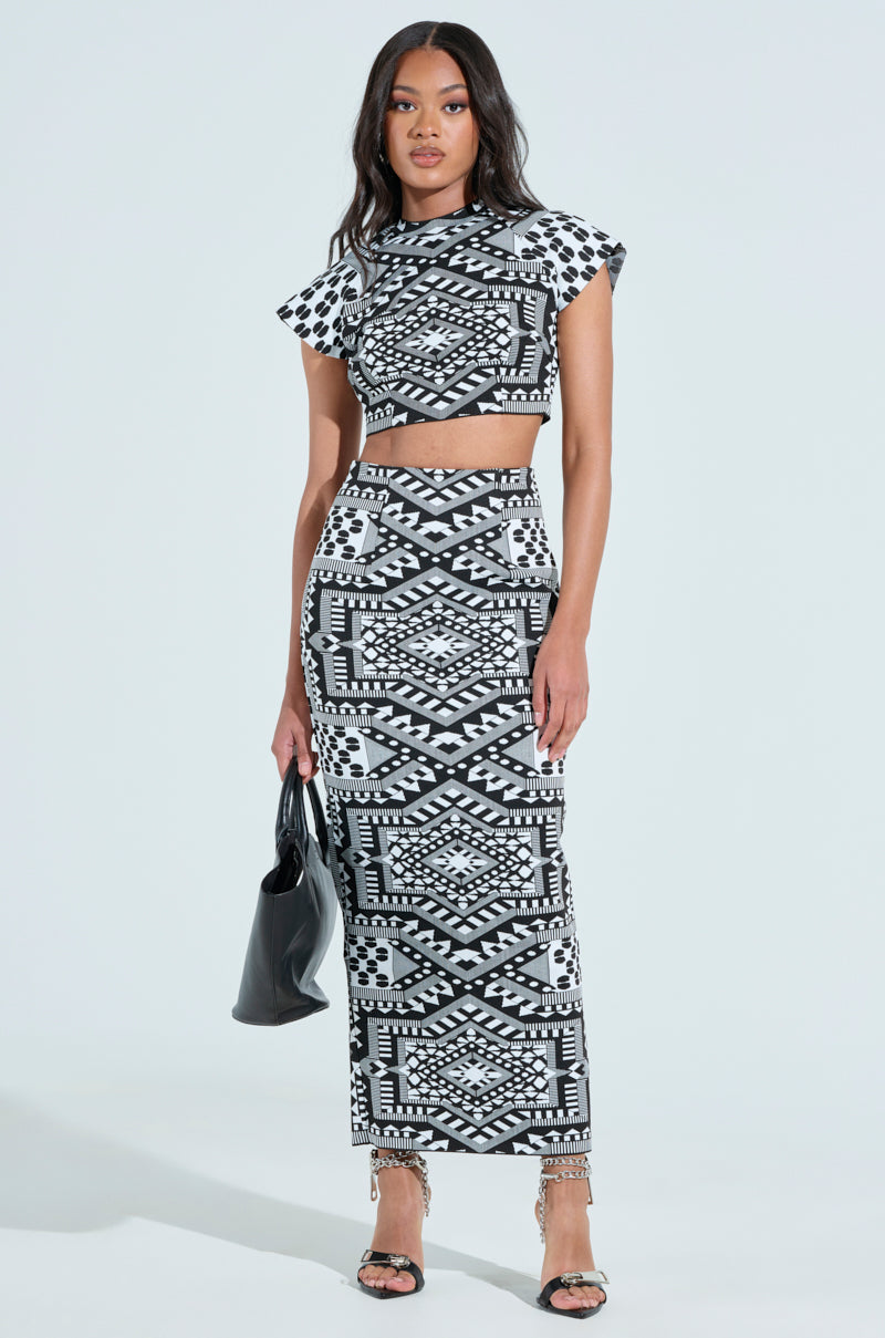 A woman poses elegantly against a light background, wearing a geometric B&W two-piece set featuring a short-sleeved, cropped top and the high-rise VACA MODE ABSTRACT MAXI SKIRT. She carries a black bag and completes her look with strappy heels.
