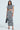 A woman poses elegantly against a light background, wearing a geometric B&W two-piece set featuring a short-sleeved, cropped top and the high-rise VACA MODE ABSTRACT MAXI SKIRT. She carries a black bag and completes her look with strappy heels.