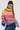 A person with long braided hair is wearing the LOOK AT ME CHUNKY MULTI COLOR KNIT SWEATER, featuring a vibrant and oversized silhouette with a rainbow striped pattern in shades of yellow, pink, blue, and purple. They pair it with a denim skirt and large hoop earrings against a plain white background.