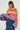 A person with long braided hair dons the LOOK AT ME CHUNKY MULTI COLOR KNIT SWEATER, showcasing a vibrant rainbow striped pattern of green, red, purple, and pink. The oversized silhouette is styled with a denim skirt and hoop earrings while they pose against a plain background.