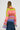 A person with braided hair is wearing the LOOK AT ME CHUNKY MULTI COLOR KNIT SWEATER in green, red, orange, and purple hues. They are facing away from the camera, showcasing an oversized silhouette while holding a white purse and pairing it with a blue denim skirt.
