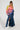 The individual is wearing the LOOK AT ME CHUNKY MULTI COLOR KNIT SWEATER featuring a rainbow striped pattern, accompanied by a long denim skirt with a slit and white knee-high boots. They have long braided hair and are posed confidently against a plain background, showcasing their oversized silhouette.