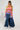 A person wearing the LOOK AT ME CHUNKY MULTI COLOR KNIT SWEATER with a rainbow striped pattern and long bell sleeves, paired with a long denim skirt featuring front slits and white ankle boots. Their hair is styled in long braids against a plain, light-colored background.