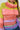 A person wearing the LOOK AT ME CHUNKY MULTI COLOR KNIT SWEATER, with its oversized design and vibrant rainbow stripes in shades of green, pink, orange, purple, and blue. Long braids cascade over the chunky knit as they stand confidently with one arm bent.