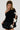 A person with braided hair wears the COQUETTE CHIC CHUNKY SWEATER IN BLACK, which boasts a stylish one-sleeve design with contrast bow appliques and an asymmetrical foldover neckline. Complemented by a black puffy skirt, they stand against a plain background, looking down with a subtle expression.