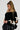A person with braided hair wears the Coquette Chic Chunky Sweater in black, featuring an asymmetrical foldover neckline adorned with contrast bow appliques, and pairs it with a sleek black skirt. They stand against a plain background, looking down with their hands gently clasped.
