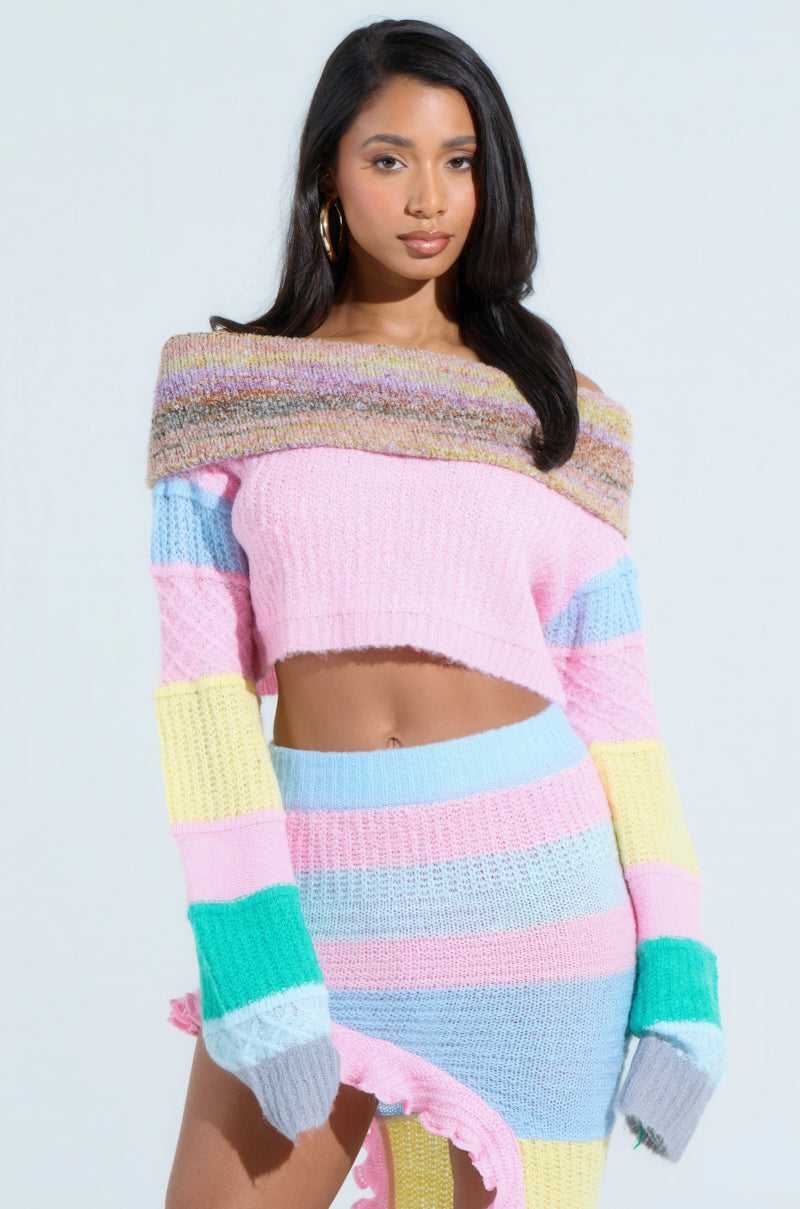 A woman stands elegantly against a light background, wearing the PRETTY LIKE ME OFF THE SHOULDER SWEATER with pastel color-blocking in pink, purple, yellow, and blue stripes. She pairs it with a matching textured skirt, complementing her long dark hair and hoop earrings.