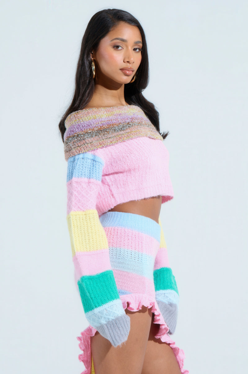 A woman poses against a light gray background in the PRETTY LIKE ME OFF THE SHOULDER SWEATER and matching skirt. The cozy pastel-colored color-block design features pink, blue, yellow, and green with a textured look. She has long hair and wears hoop earrings.