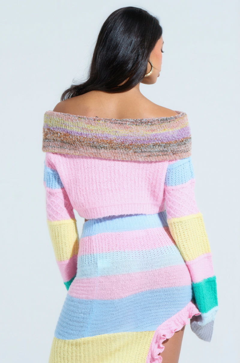 A woman with long dark hair faces away, wearing the PRETTY LIKE ME OFF THE SHOULDER SWEATER. It has pastel stripes of pink, blue, yellow, and green in a color-block design. The dress features a high-low ruffled hemline and is paired with large hoop earrings.