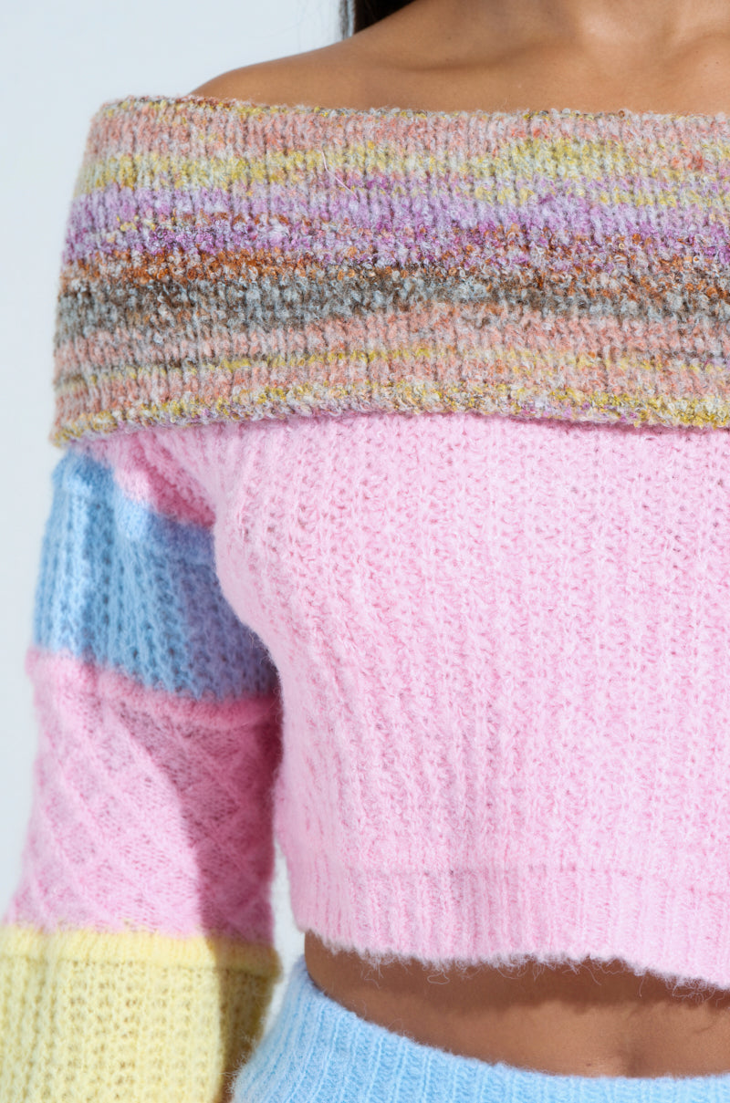 Wearing the PRETTY LIKE ME OFF THE SHOULDER SWEATER, a person enjoys cozy knit comfort with pastel pink, blue, and yellow in a color-block pattern. The sweater includes multicolored stripes at the neckline, long sleeves, and a cropped fit against a light-colored background.