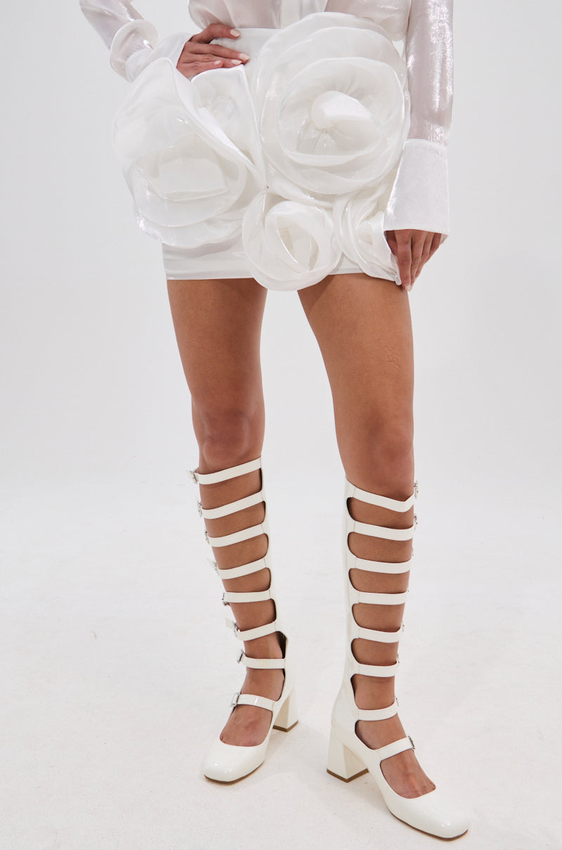 Wearing the PRETTY HOT MINI SKIRT, a white high-rise mini with bold rose appliques, and white knee-high gladiator chunky-heeled shoes, a person stands against a plain white background. 3