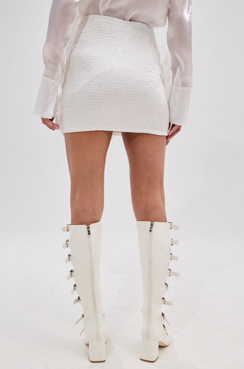 Facing away, a person wears the PRETTY HOT MINI SKIRT—a textured white high-rise skirt—styled with a sheer long-sleeve blouse and knee-high white boots featuring chunky heels and side buckles. The background is plain white. 5