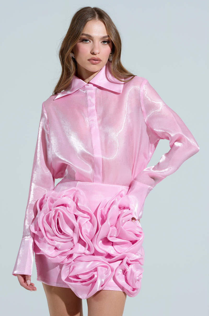 A woman poses in the PRETTY HOT BUTTON DOWN TOP, a shiny pink blouse with snap button closures, and a matching skirt featuring large textured floral designs. She has long, wavy hair and stands against a simple light gray background.