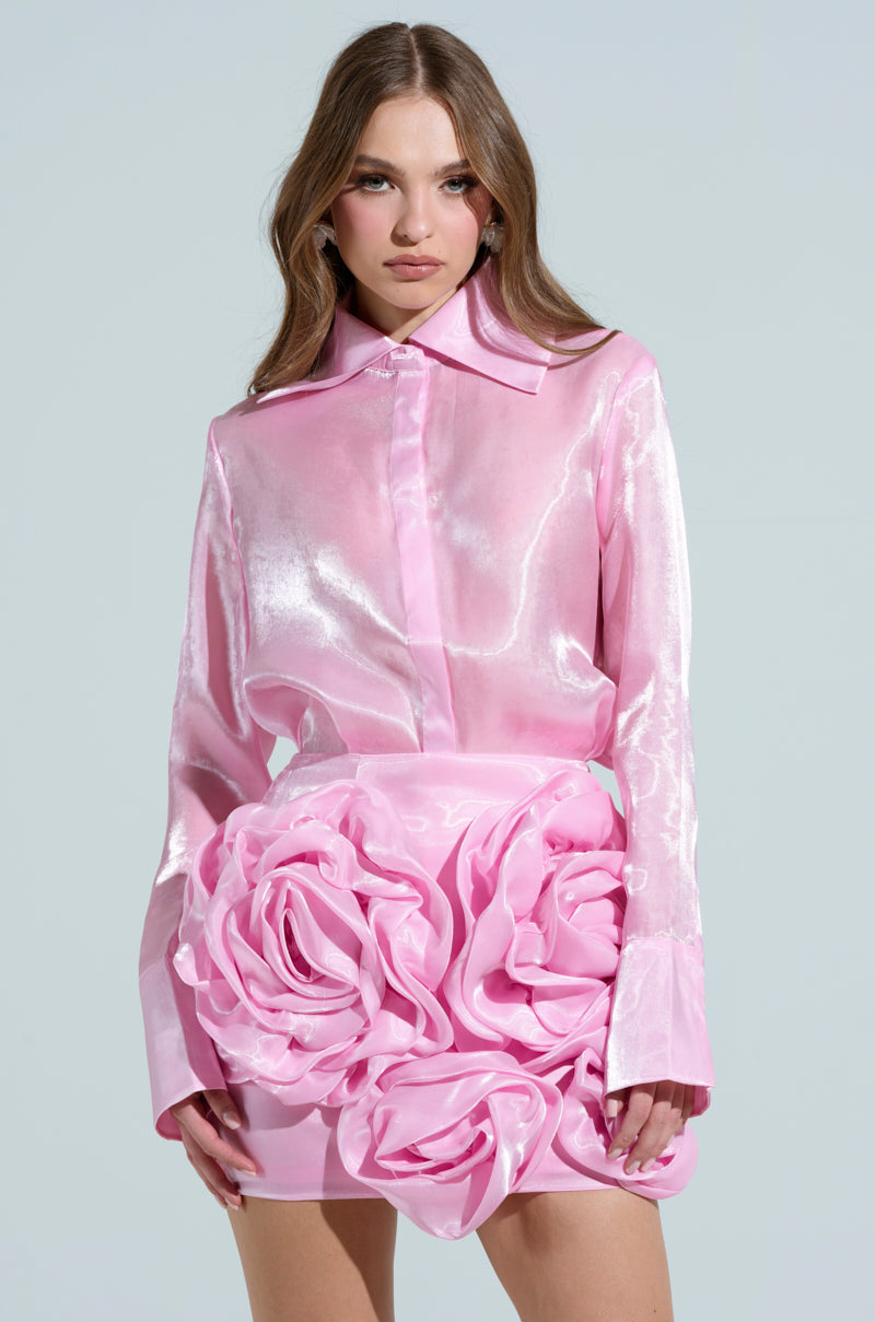 A person poses in the PRETTY HOT BUTTON DOWN TOP, a shiny pink blouse with snap buttons, matched with a pink skirt featuring large fabric roses. They have long wavy hair and a neutral expression against a plain light gray background.