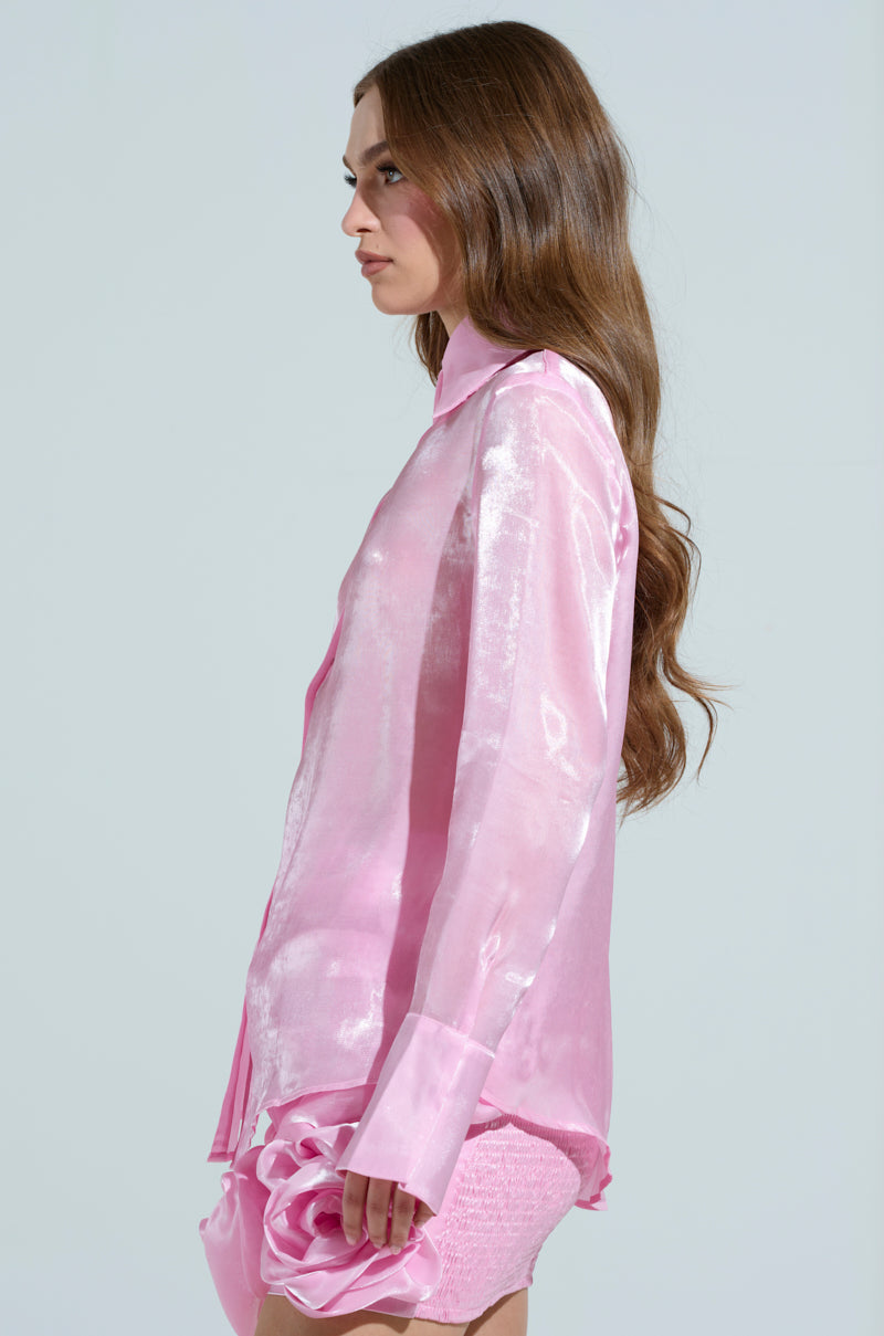 A person with long brown hair is wearing the PRETTY HOT BUTTON DOWN TOP, a shiny pink blouse with snap button closures, and a matching pink skirt with a textured floral design featuring an elegant longline silhouette. The backdrop is plain gray.