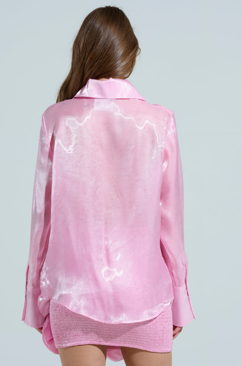 A woman with long brown hair faces away, wearing the PRETTY HOT BUTTON DOWN TOP, a shiny pink satin blouse with snap button closures and a textured skirt. The longline silhouette complements the simple backdrop of plain light gray.