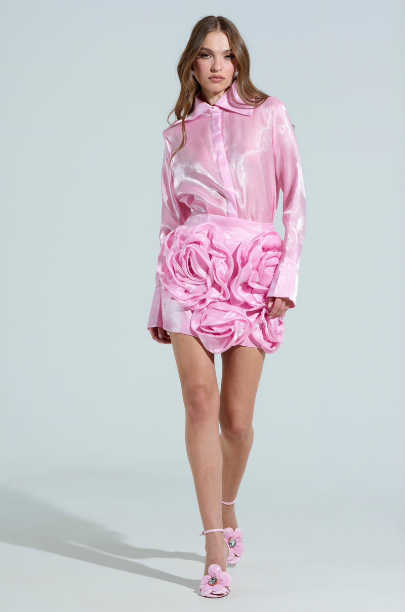 A model showcases the PRETTY HOT BUTTON DOWN TOP, a shiny pink blouse with snap closures, pairing it with a rose-patterned skirt. She completes the look with flower-accented pink shoes against a plain, light backdrop.
