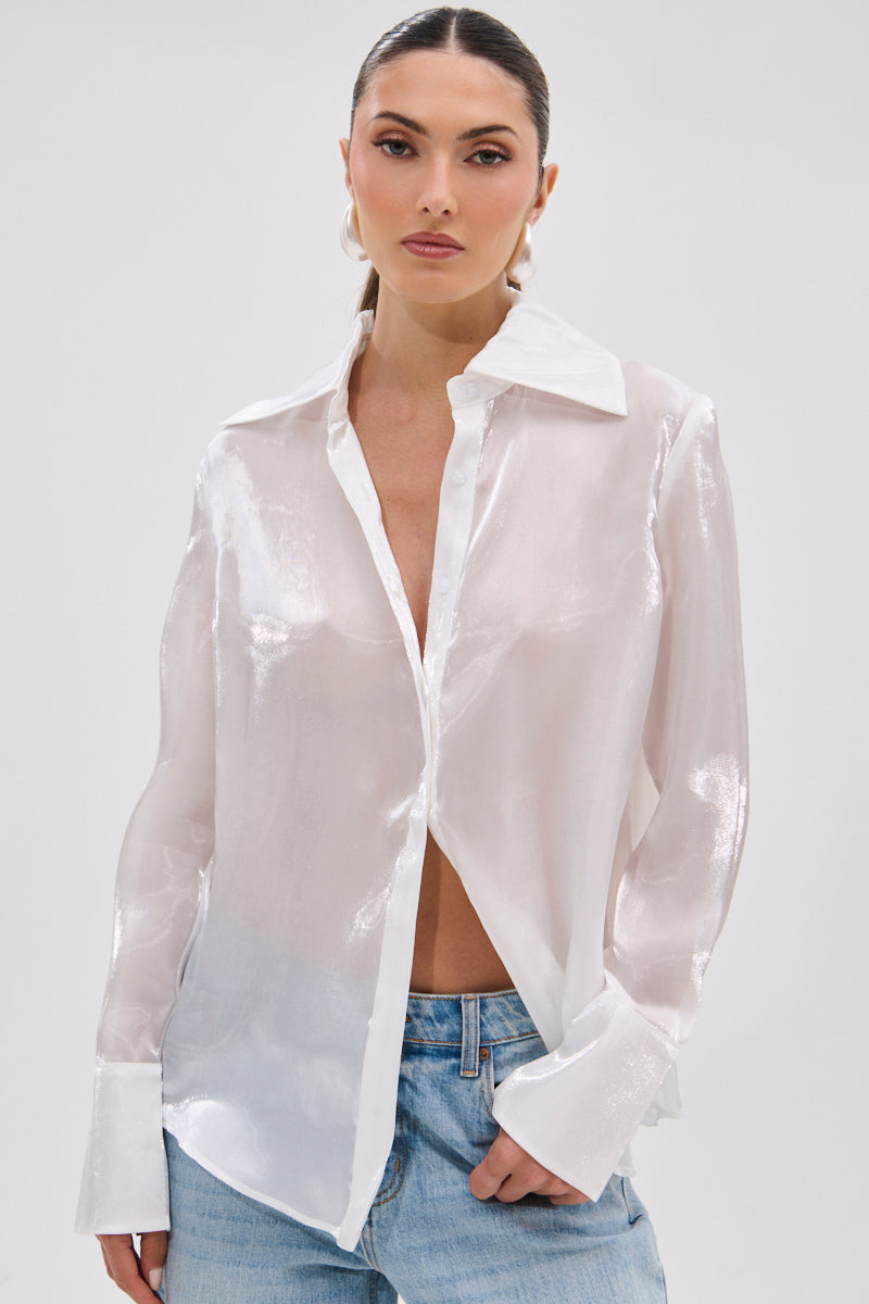 A woman with slicked-back hair stands against a plain light background, wearing the PRETTY HOT BUTTON DOWN—an elegant semi-sheer, lustrous white blouse—styled with blue jeans.