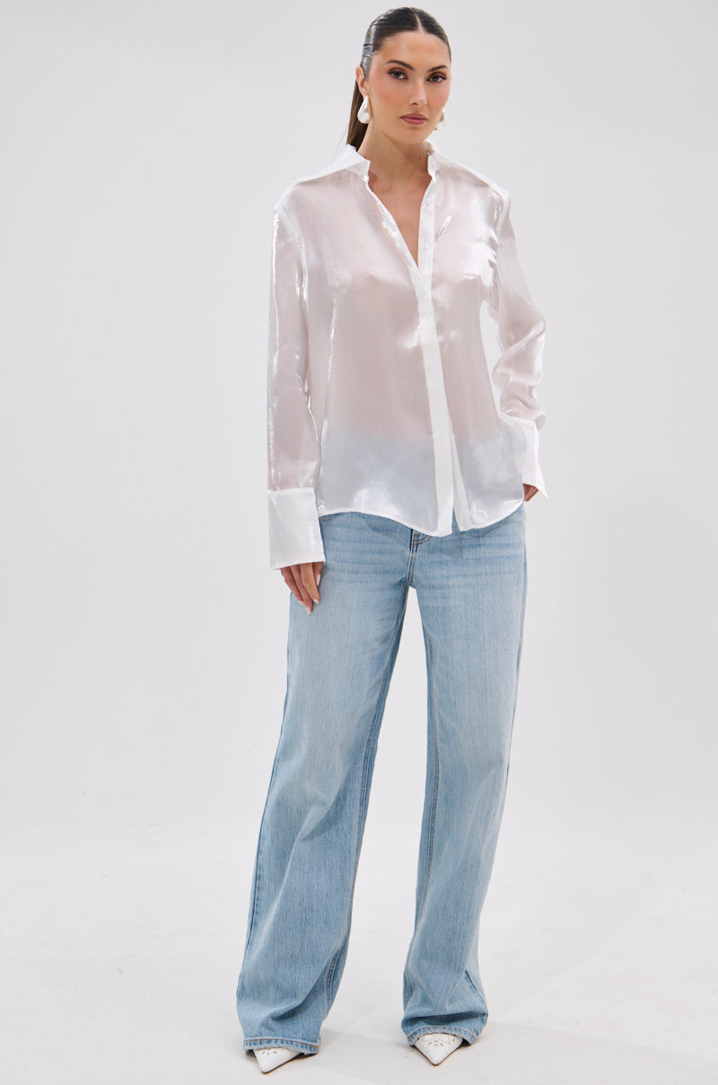 A woman stands against a plain background wearing the PRETTY HOT BUTTON DOWN, a lustrous white long-sleeve blouse, paired with light blue wide-leg jeans and white pointed-toe shoes. Her hair is pulled back.