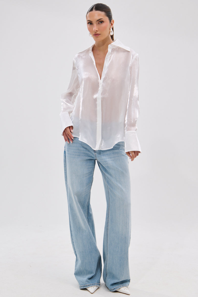 A woman stands against a plain background wearing the PRETTY HOT BUTTON DOWN—a semi-sheer, lustrous blouse with wide cuffs—paired with light blue wide-leg jeans, white pointed-toe shoes, and large hoop earrings. Her hair is pulled back for a chic look.