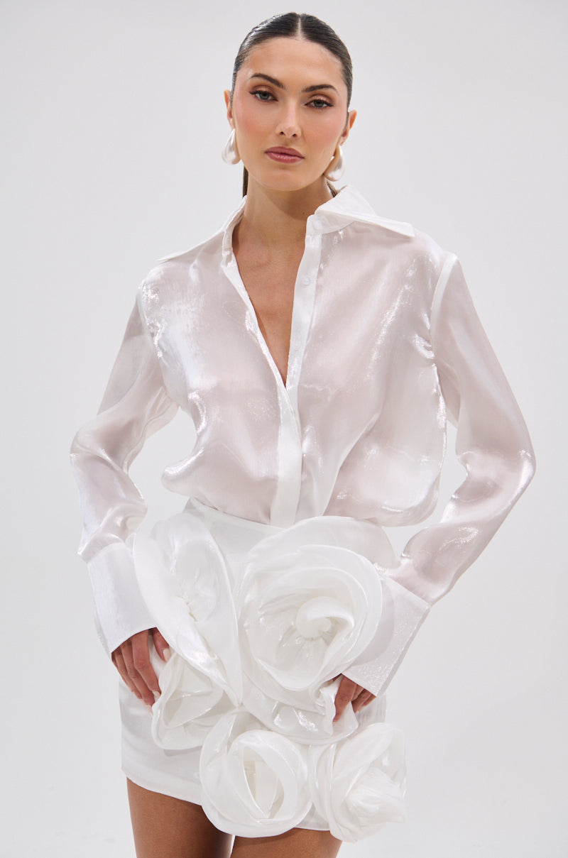 A person models the PRETTY HOT BUTTON DOWN, a semi-sheer, lustrous white blouse paired with a white skirt featuring large sculpted fabric flowers, posing against a plain light background.