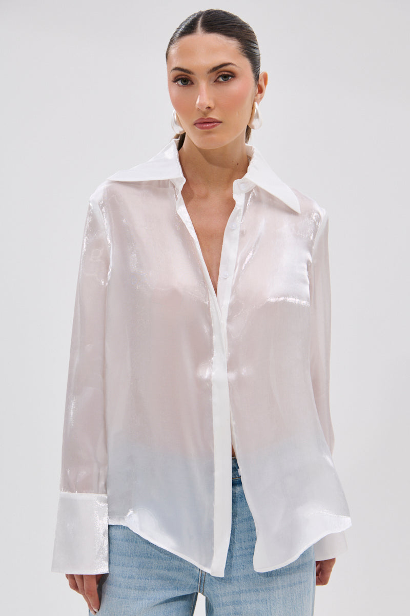 A woman models the PRETTY HOT BUTTON DOWN, a lustrous semi-sheer blouse with wide cuffs and collar, paired with light blue jeans. She stands against a plain white background, hair pulled back and expression neutral.