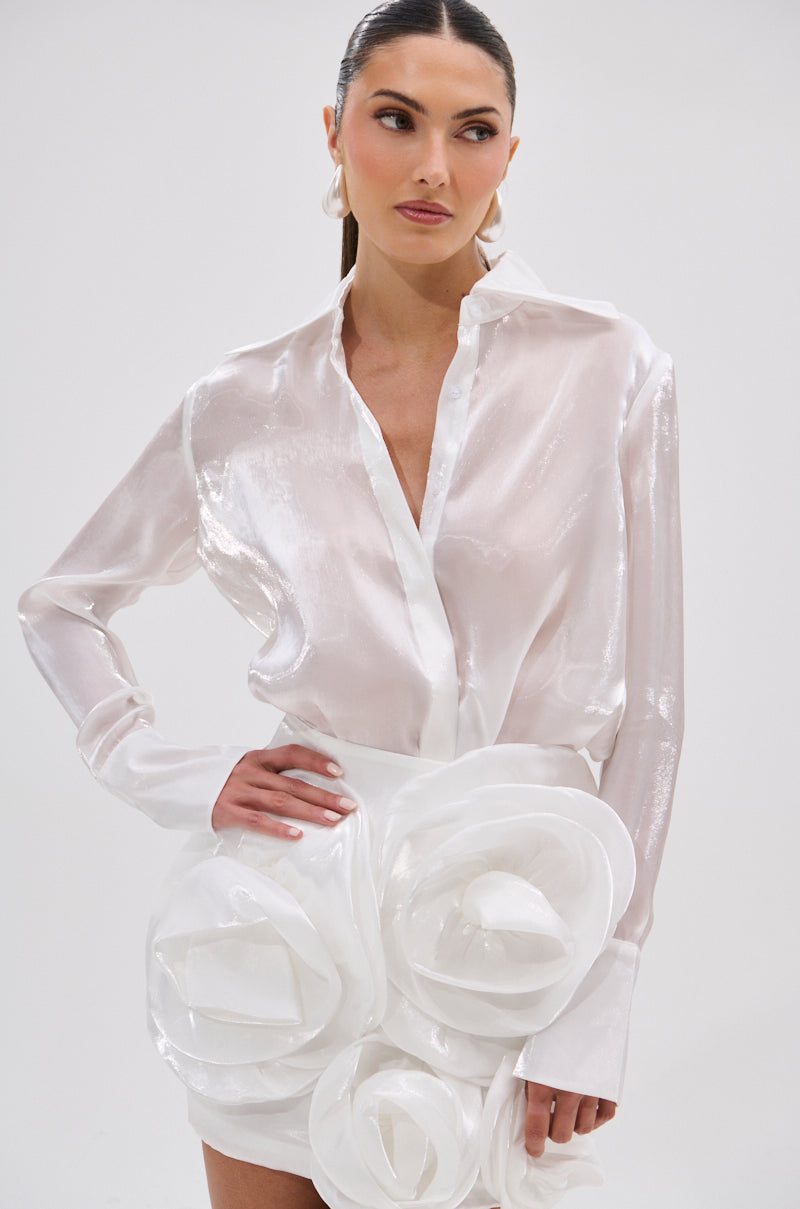 A woman with long dark hair in a sleek ponytail confidently models the PRETTY HOT BUTTON DOWN, a chic semi-sheer white blouse paired with a white skirt featuring large sculpted fabric roses, against a plain background.