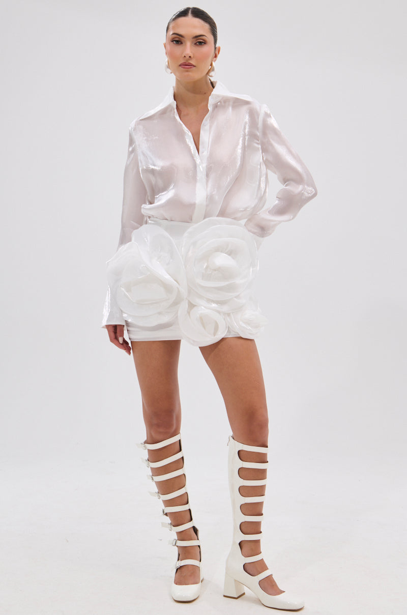A woman poses in the PRETTY HOT BUTTON DOWN, a lustrous, semi-sheer white blouse, paired with a rose-adorned white mini skirt and knee-high block heel gladiator boots against a plain white background.