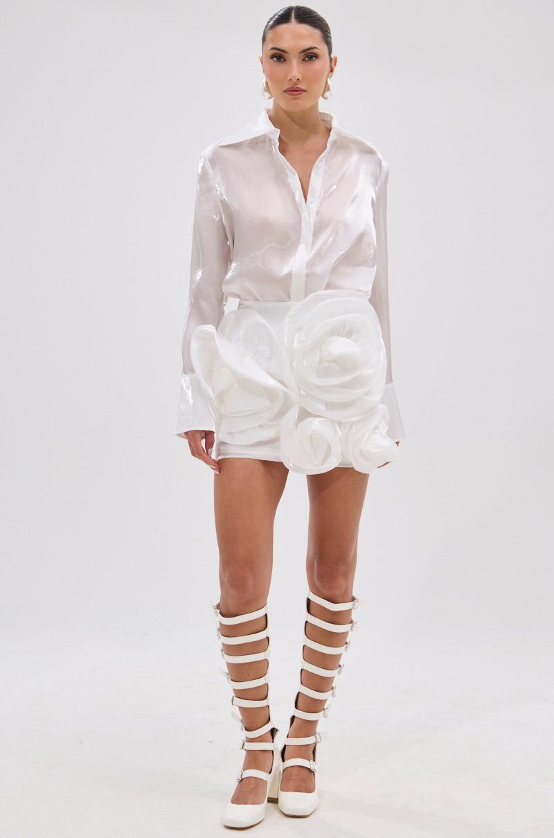 A woman stands against a plain background in the PRETTY HOT BUTTON DOWN, a chic semi-sheer white blouse paired with a lustrous mini skirt with sculptural floral details and knee-high white strappy sandals. Her hair is slicked back and her expression is neutral.